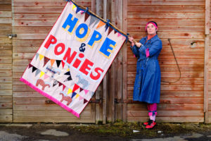 A woman in a blue denim dress and wearing pink-fringed headband and socks stands in front of a lapped wooden wall holding a large banner with the words ‘Hope and Ponies’