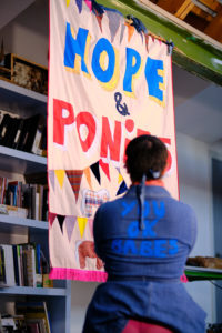 A woman in a blue denim dress sits in front, facing a large banner with the words ‘Hope and Ponies"