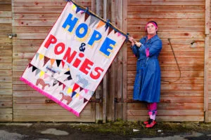 A woman in a blue denim dress and wearing pink-fringed headband and socks stands in front of a lapped wooden wall holding a large banner with the words ‘Hope and Ponies’