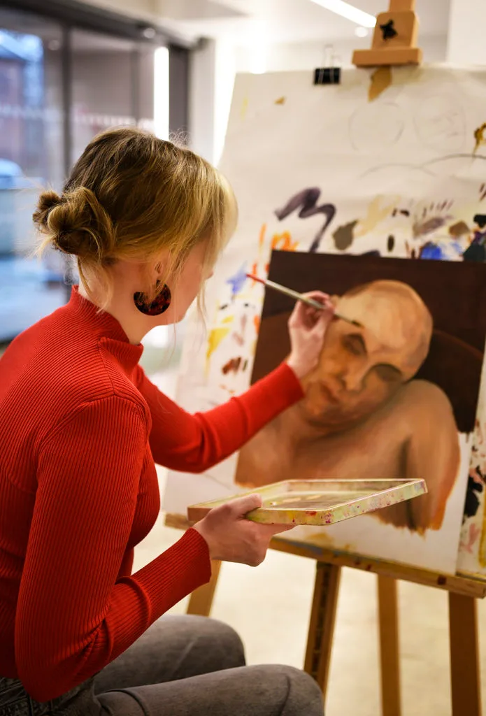 A woman wearing a red jumper sat at an easel painting a portrait.